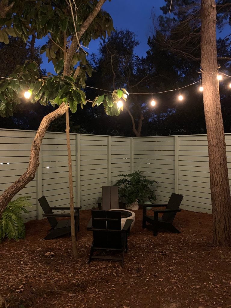firepit-area-at-night-768x1024-1 Nighttime view of fenced side yard with black Adirondack chairs around fire pit